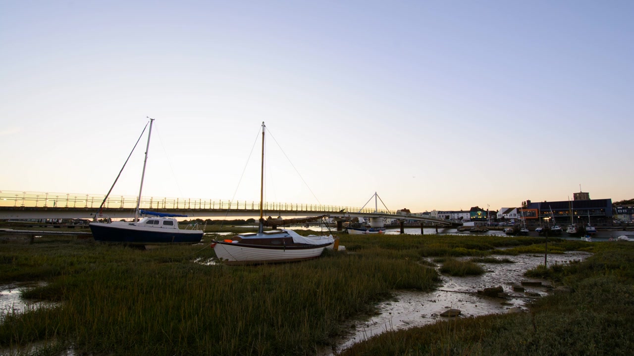 Sunset over a coastal town with boats in a river, transitioning from golden hour to darkness as town buildings and street lights gradually illuminate x2 speed