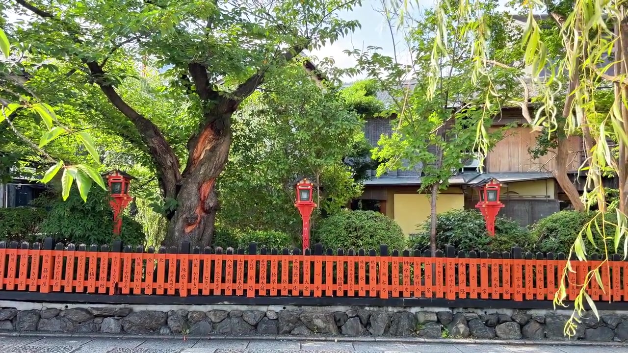 Des lanternes traditionnelles en bois et une clôture orange bordent une rue calme avec une végétation luxuriante.