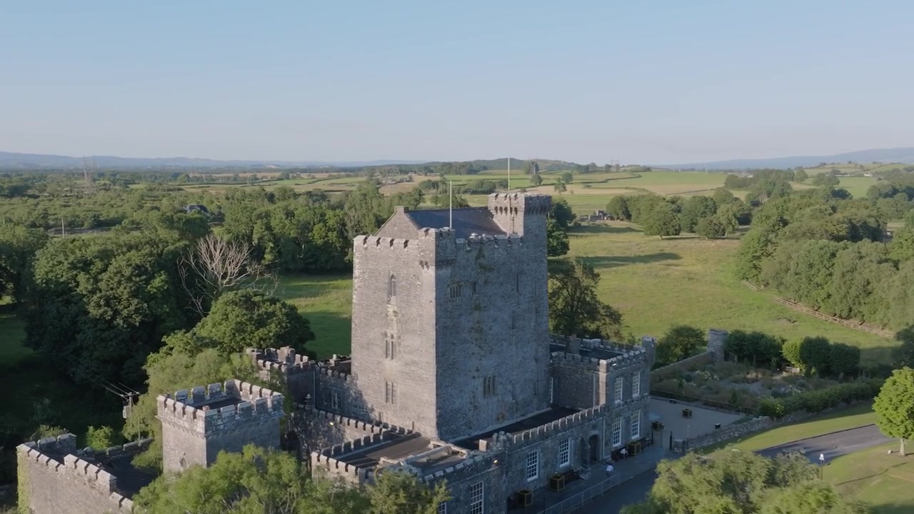 Hrad Knappogue se tyčí uprostřed širokých trávníků a stromů, zatímco se letecký záběr oddaluje, aby odhalil okolní irskou zemědělskou krajinu.