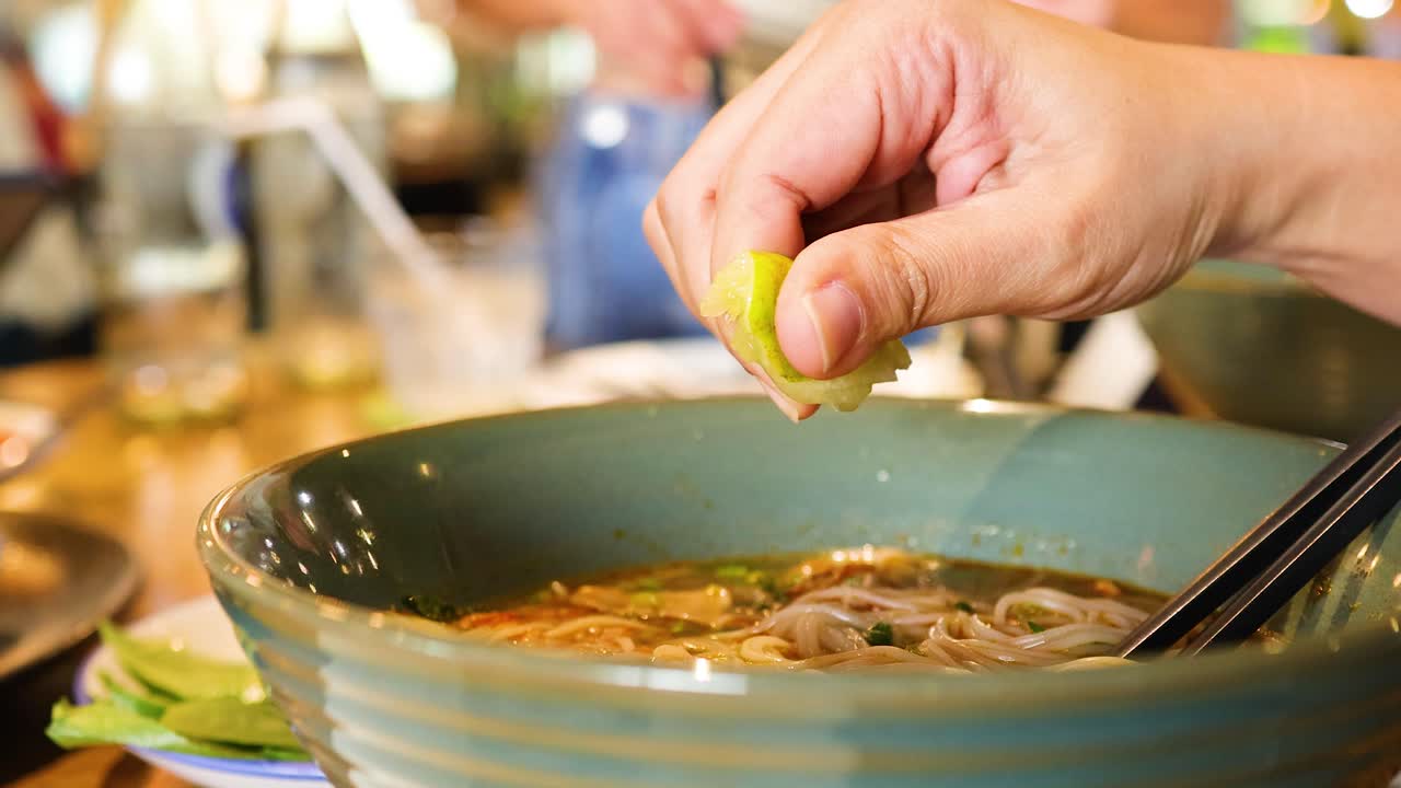 A hand squeezes lime over a bowl of pho in a bustling Bangkok restaurant, enhancing the dish's flavor