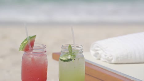 Two-tropical-drinks-in-mason-jars,-beside-a-beach-chair-with-the-ocean-shore-in-the-background