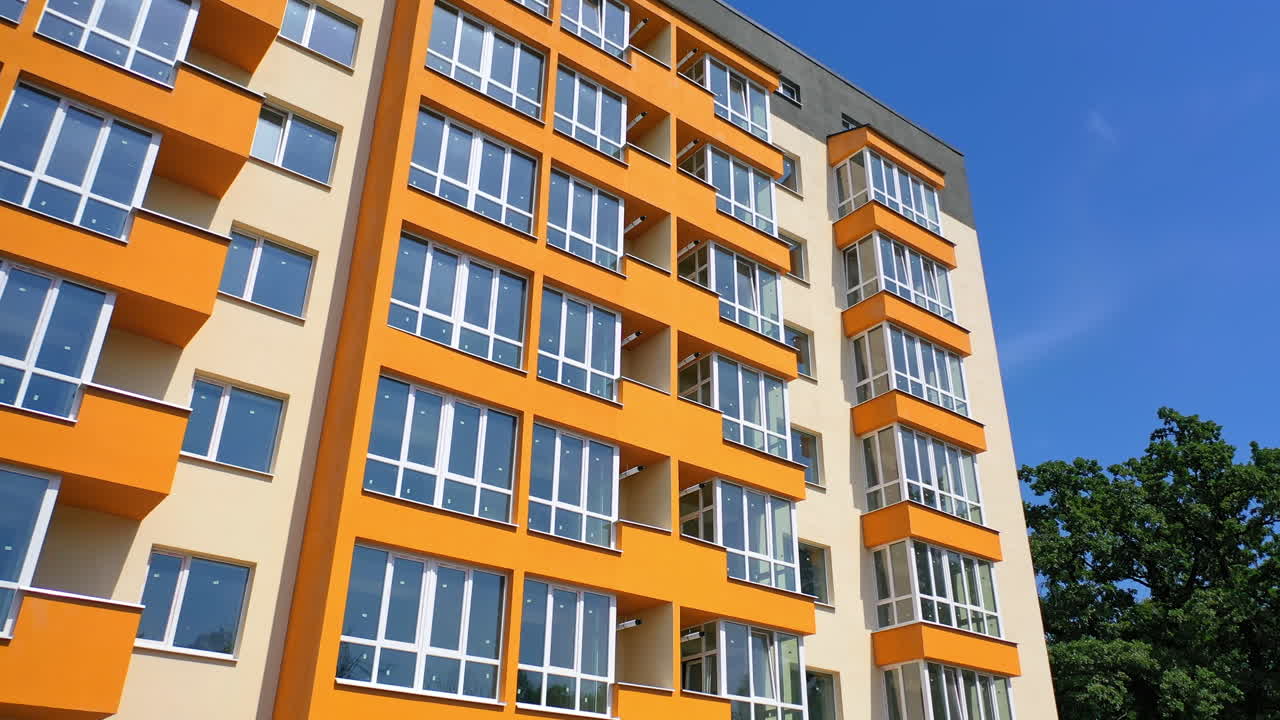 Beautiful high-rise building. Modern block of flats with large balconies on blue sky background in summer. Apartment building of colorful walls. Camera moves down.
