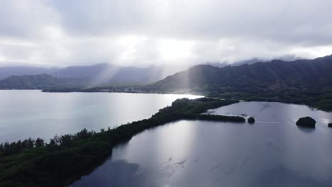 Una-Impresionante-Vista-Aérea-Que-Captura-Los-Rayos-Del-Sol-Atravesando-Las-Nubes-Sobre-Las-Montañas-Koolau,-Iluminando-La-Serena-Bahía-Y-La-Exuberante-Vegetación-Debajo.