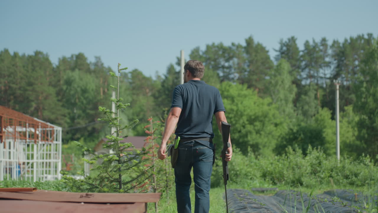 Man walking with rifle through crop rows surrounded by plants and metal frame structure, summer rural scene showing cautious patrol and protection of garden with forest backdrop