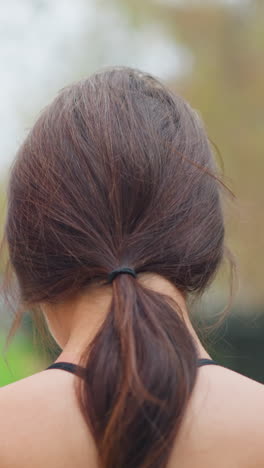 back view of young woman performing neck stretch outdoors, making neck flexible with blurred background of trees and iron fence in park, promoting relaxation and flexibility