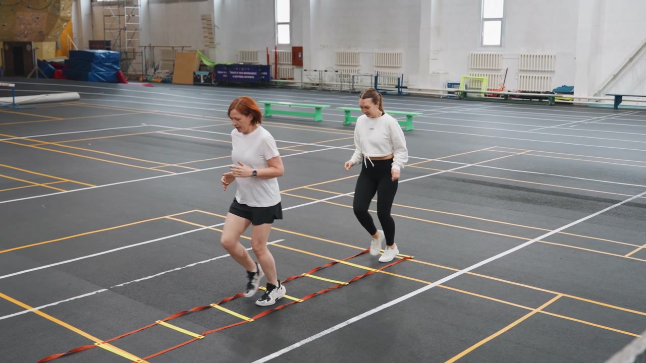 Two white women practicing badminton footwork ladder drills on indoor court, coach demonstrating quick steps while teammate follows, marked court lines and colored cones, focused expressions