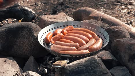 un hombre pone salchichas a la parrilla en una sartén sobre una fogata