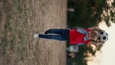 Vertical-video-A-little-blond-boy-in-a-red-T-shirt-throws-a-soccer-ball-on-the-ground-and-hits-it-with-his-foot-in-the-park.-Happy-little-boy-having-fun-and-active-time-in-the-park