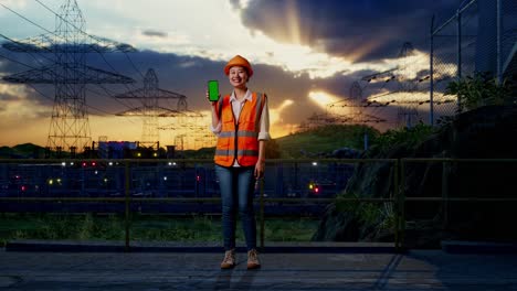 full body of asian female engineer with safety helmet smiling and showing green screen smartphone to the camera near high voltage tower
