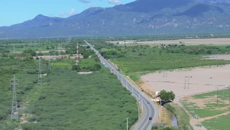 Aerial-flyover-CARRETERA-SANCHEZ-Highway-in-Azua-between-green-fields-and-mountain-range-in-background---Cars-driving-on-road