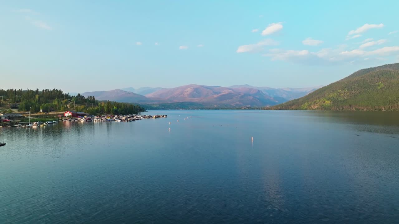 Drone flies over still lake surface with pine shoreline and mountain backdrop at Grand Lake Colorado, soft daylight