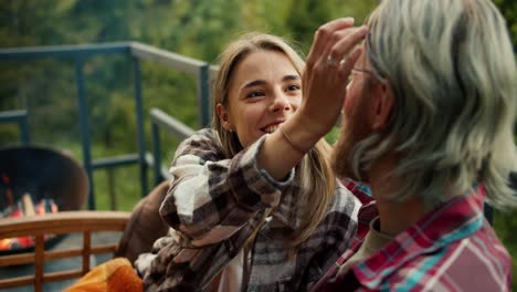 a blonde girl examines and strokes her blonde boyfriend in glasses, sitting on the sofa of a country house overlooking the mountains and a lush coniferous forest