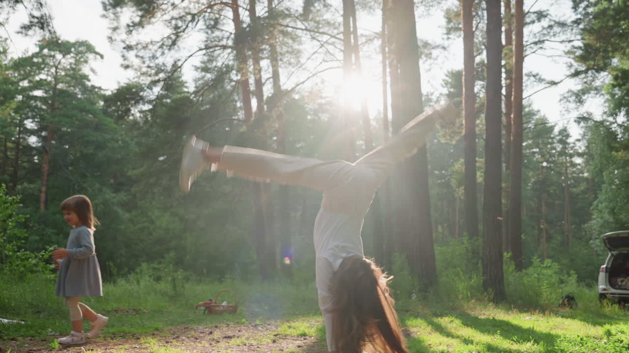 Jovencita haciendo una enérgica rueda lateral en una esterilla de picnic en un bosque soleado mientras su hermana pequeña la observa jugando cerca, rodeada de altos árboles y cálida luz solar