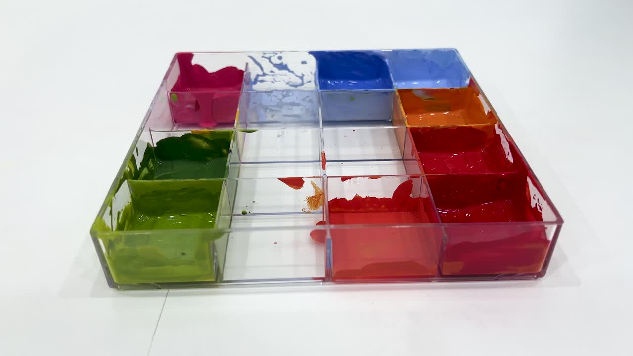 A transparent plastic tray with poster paints is gradually emptied, showing red, green, blue, and orange colors under bright, even lighting with a static overhead camera
