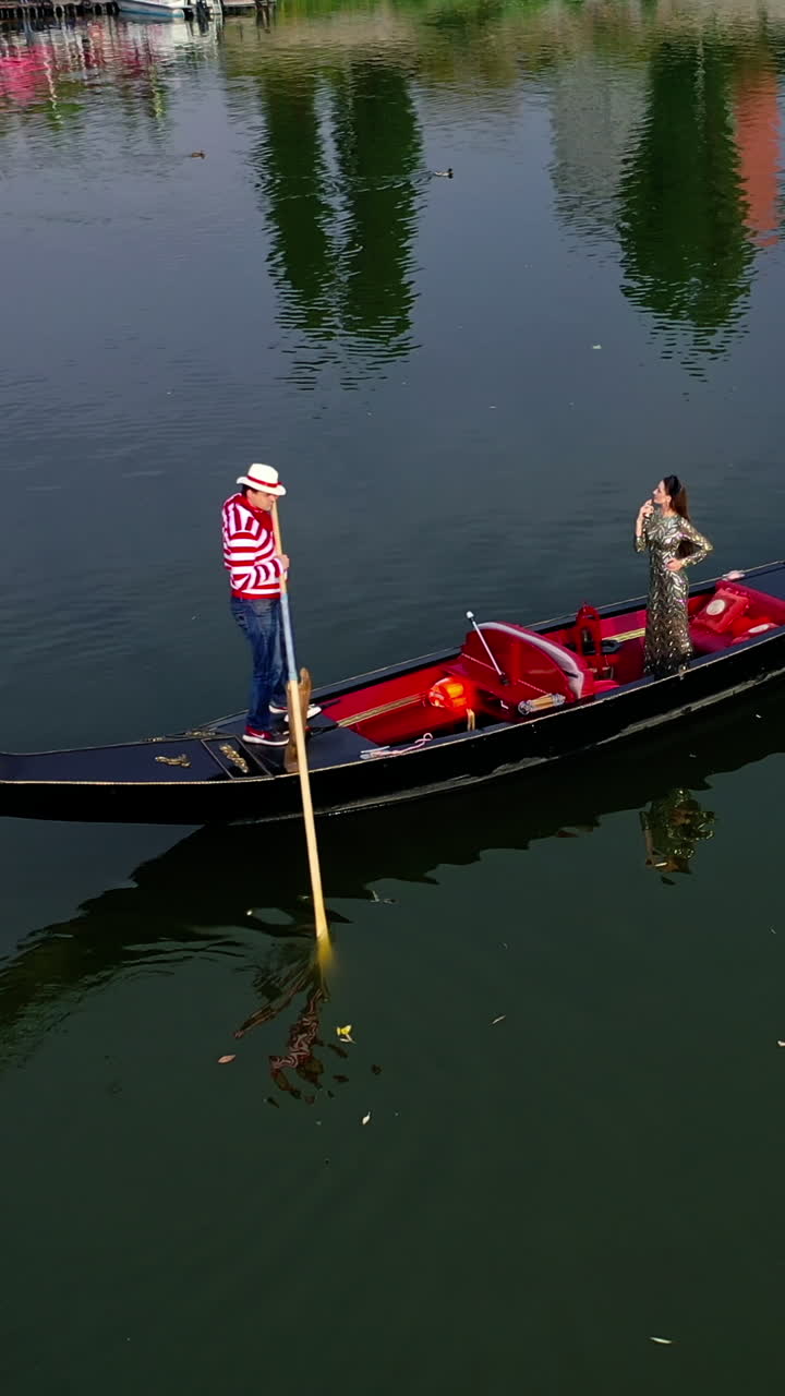 Gondolier riding a boat with a woman. Beautiful lady travelling in gondola along the river at sunset. Aerial view. Camera motion around. Vertical video