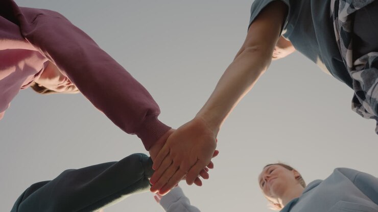Visuale dal basso di persone con felpe col cappuccio che uniscono le mani come membri di un team, con la luce del sole che crea un effetto luminoso intorno alle loro mani, a simboleggiare unità, lavoro di squadra e collaborazione