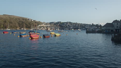 Small-Colorful-Boats-Moored-Offshore-In-Fowey-Harbour-With-View-Of-Polruan-Village-In-The-Background-In-South-Cornwall,-England---Drone-Pull-Back-Shot