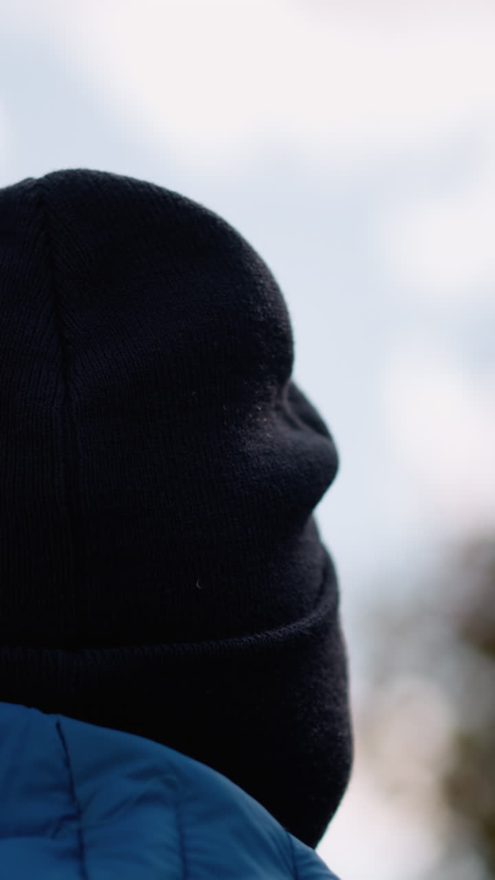 vista de la cabeza trasera de un individuo que lleva un gorro negro y una chaqueta azul, mirando al aire libre, el fondo muestra un cielo azul suave y un follaje de otoño borroso