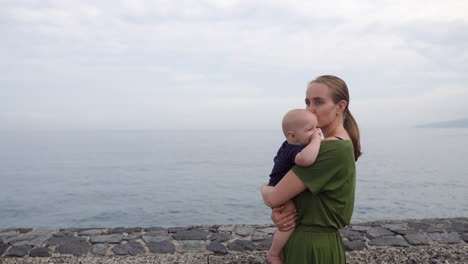 A-young-mother-strolls-with-her-baby-son-along-the-waterfront-near-the-ocean,-on-an-ancient-European-square,-watching-the-waves-and-sharing-smiles