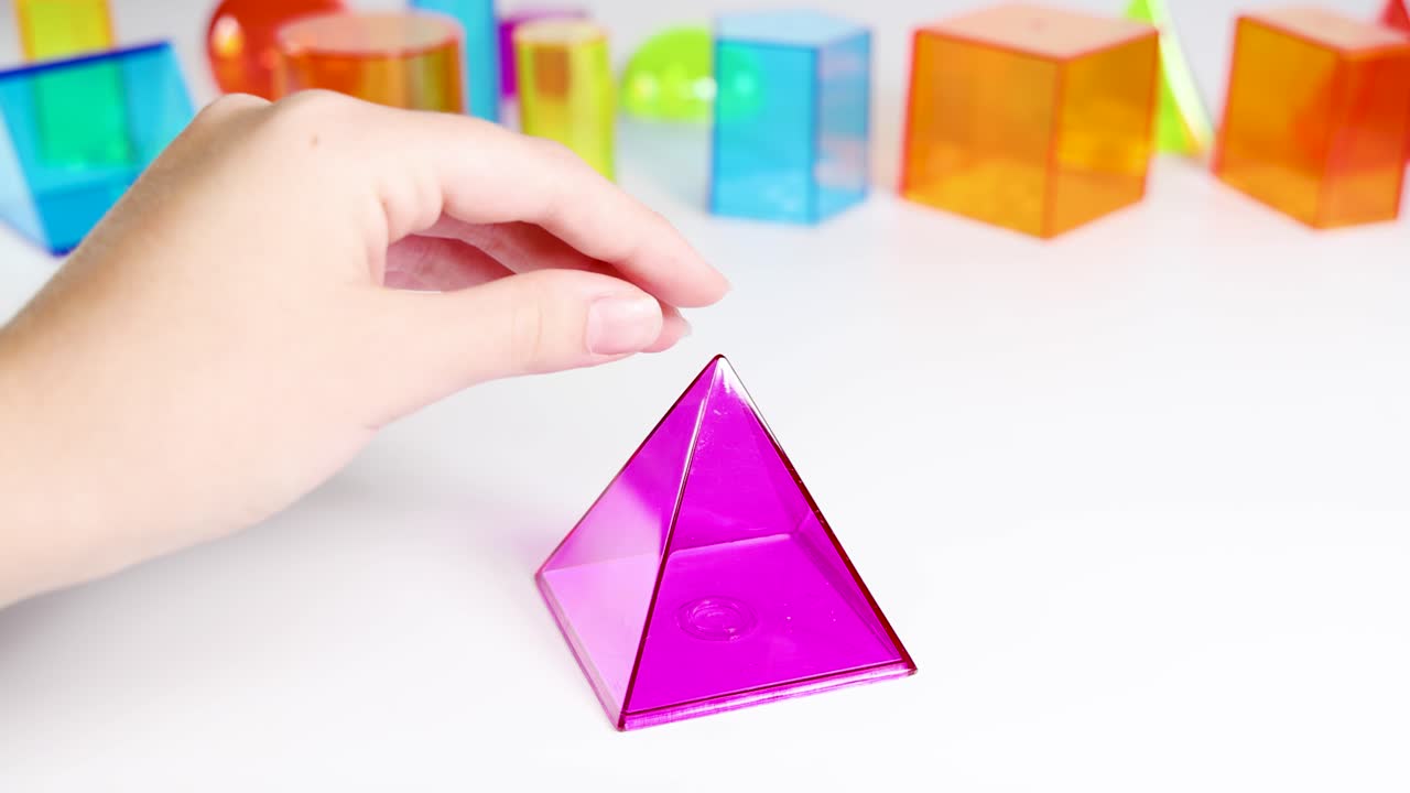 Hands manipulate colorful 3D geometric shapes on a white surface, highlighting educational exploration in bright lighting