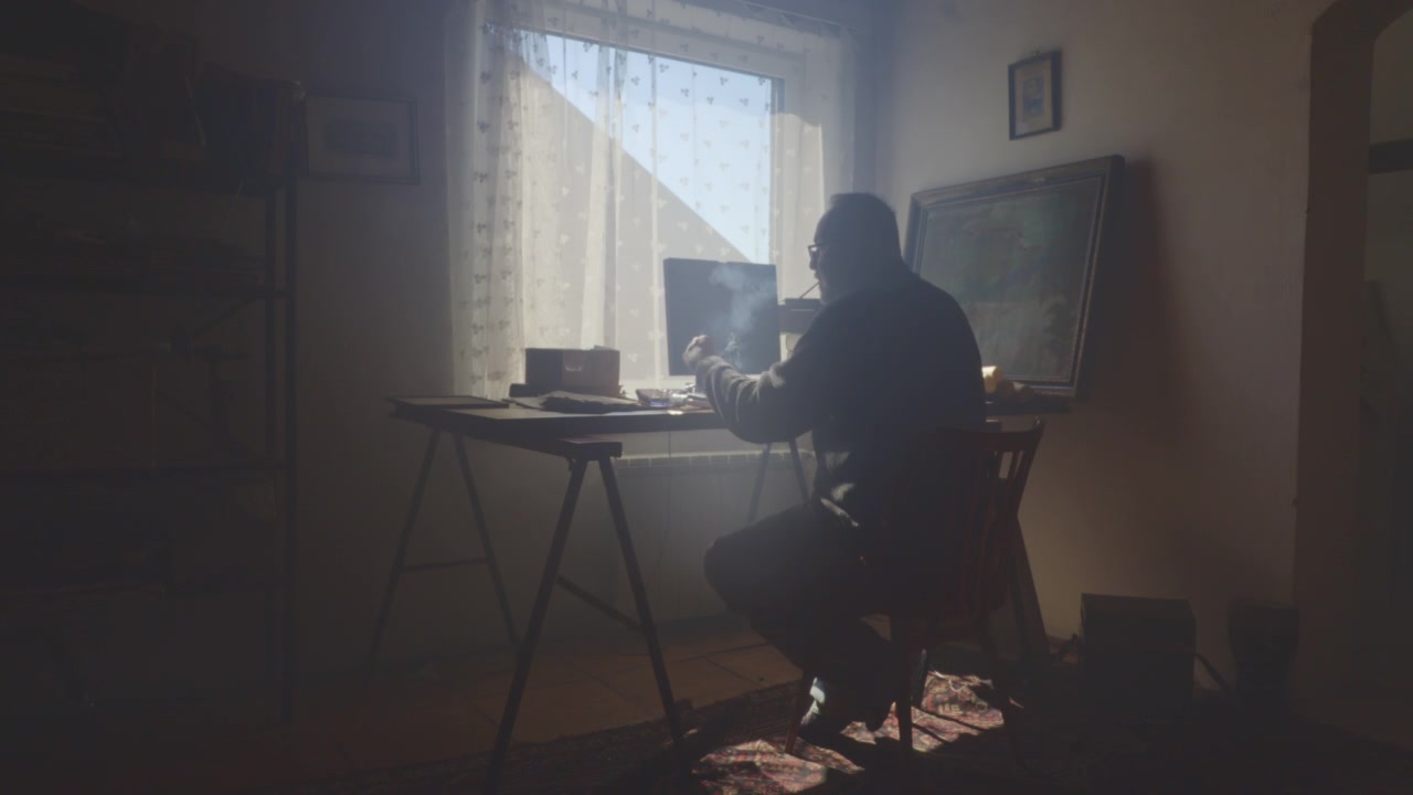 Un uomo solitario siede fumando a una scrivania con computer mentre la luce del sole entra da dietro. Filmato suggestivo che trasmette isolamento, stress, esaurimento, contemplazione e solitudine moderna