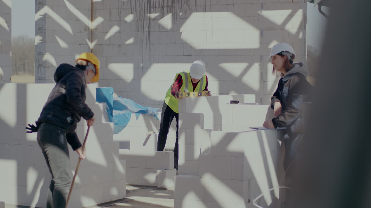 Panning shot of female worker brooming by supervisor writing on clipboard near male coworker examining wall with level at construction site