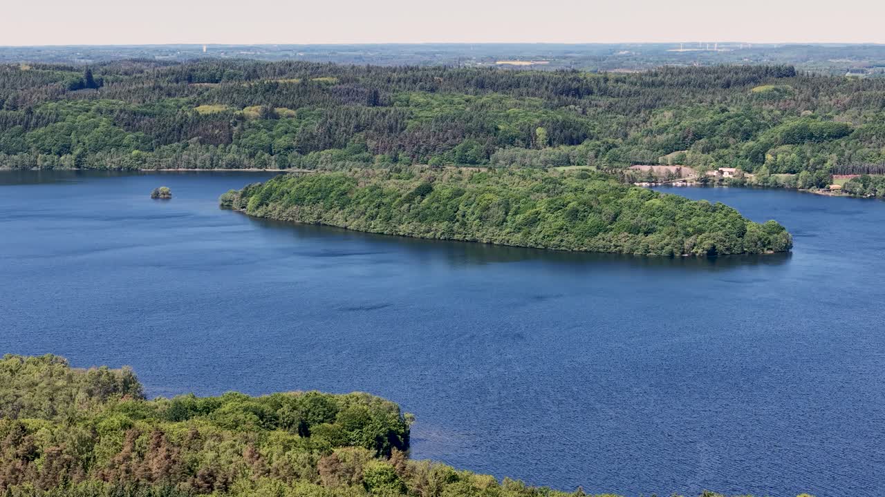 Drone footage showcasing a small forested island in a calm blue lake in Denmark, surrounded by shimmering water and distant hills under a pastel sunset sky