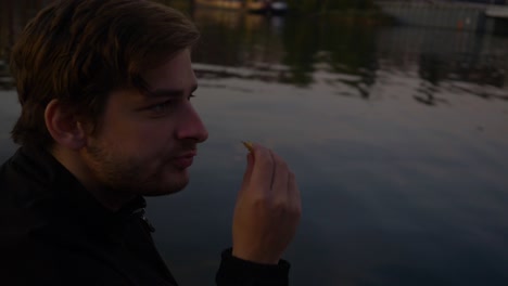 Video of a man nodding eating potato chips contently on the edge of a pier over a relfective lake during the evening
