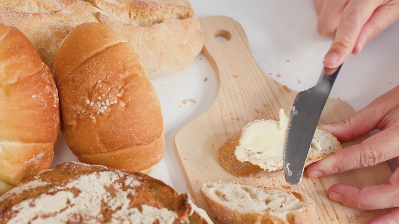 Close-up overhead shot of hands spreading creamy butter onto crusty bread on wooden board.
