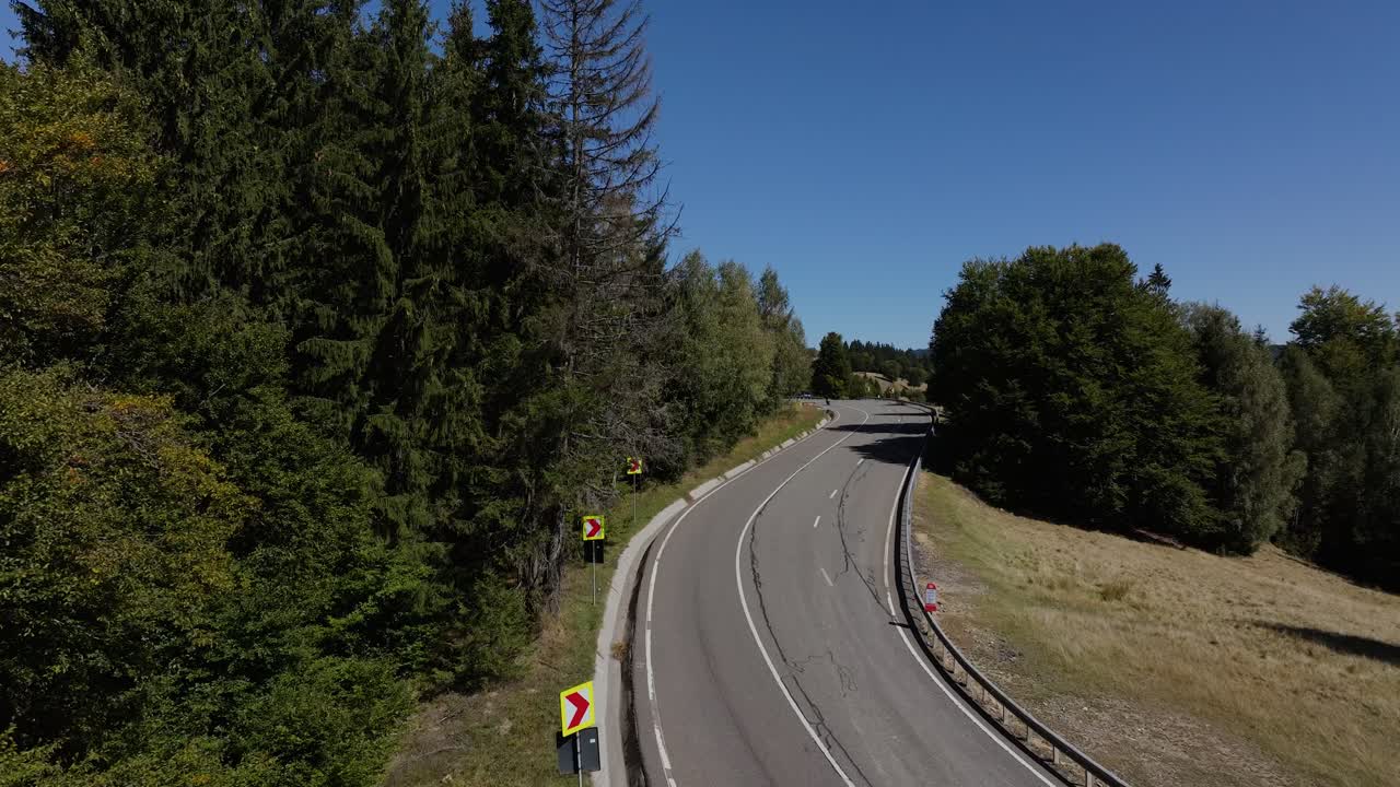Motorcyclist navigates the curving mountain road surrounded by dense forests and open grassland under a clear blue sky in bright daylight