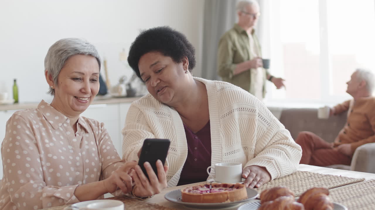 Medium close-up POV of senior Asian and African women smiling, sitting at table in foreground of blurred friends, using smartphone, talking