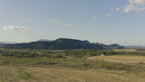 Landscape-aerial-shot-of-the-countryside-road-with-moving-cars-passing-through-the-fields-with-haystacks-and-mountain-ridge-in-the-background-on-a-clear-sunny-day,-Russia,-far-East