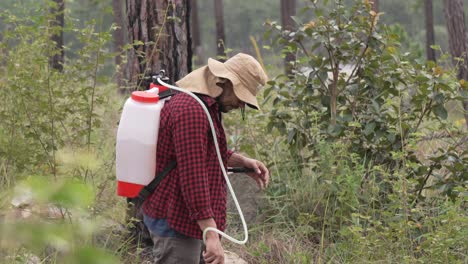 Medium-shot-of-a-man-forest-ranger-working-with-a-backpack-sprayer-in-the-forest,-he-wears-a-red-shirt-and-a-hat