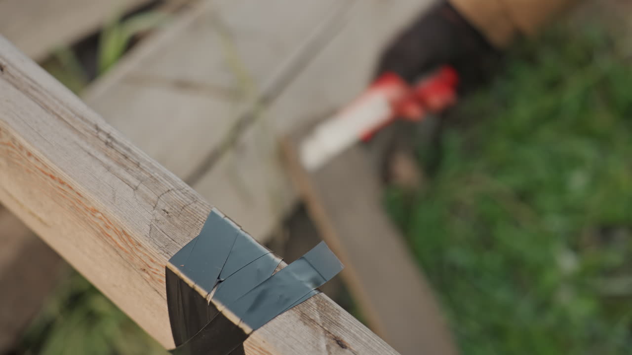 Primo piano della riparazione di una tavola di legno con nastro adesivo, mani guantate che tagliano e levigano la giunzione del nastro con un taglierino rosso, ambiente di costruzione esterno, attento restauro e manutenzione di legname stagionato, esperto.