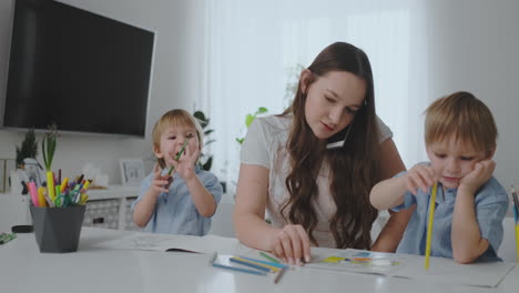 a young mother with two children talking on a mobile phone draws with a pencil and helps children draw with colored pencils