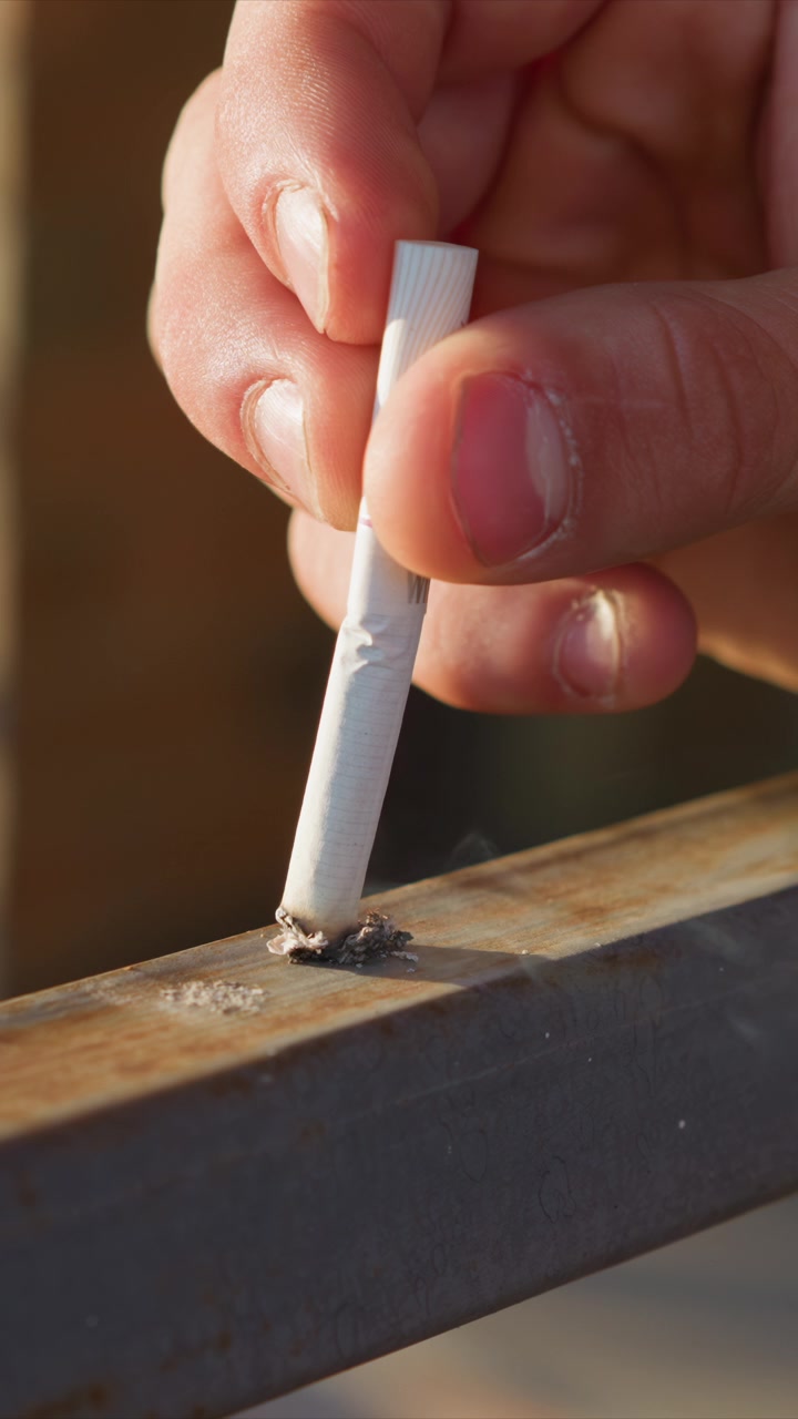 Primer plano de una mano sosteniendo un cigarrillo apagado sobre metal, bajándolo repetidamente para dejar ceniza. El enfoque se mantiene en la interacción entre los dedos y los materiales para fumar contra texturas industriales.