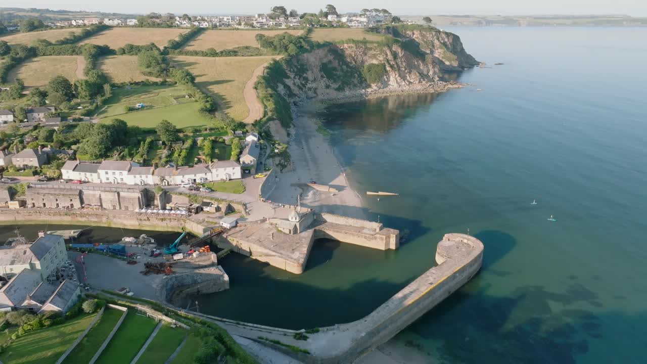 Port village with harbour walls, sea cliffs and beaches with camera reverse over green fields and trees. Charlestown, Cornwall, UK.