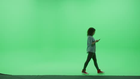 green screen chroma key studio: young girl wearing stylish casual clothes uses smartphone, browses through internet, chats on social media walks across room left to right. side view camera shot