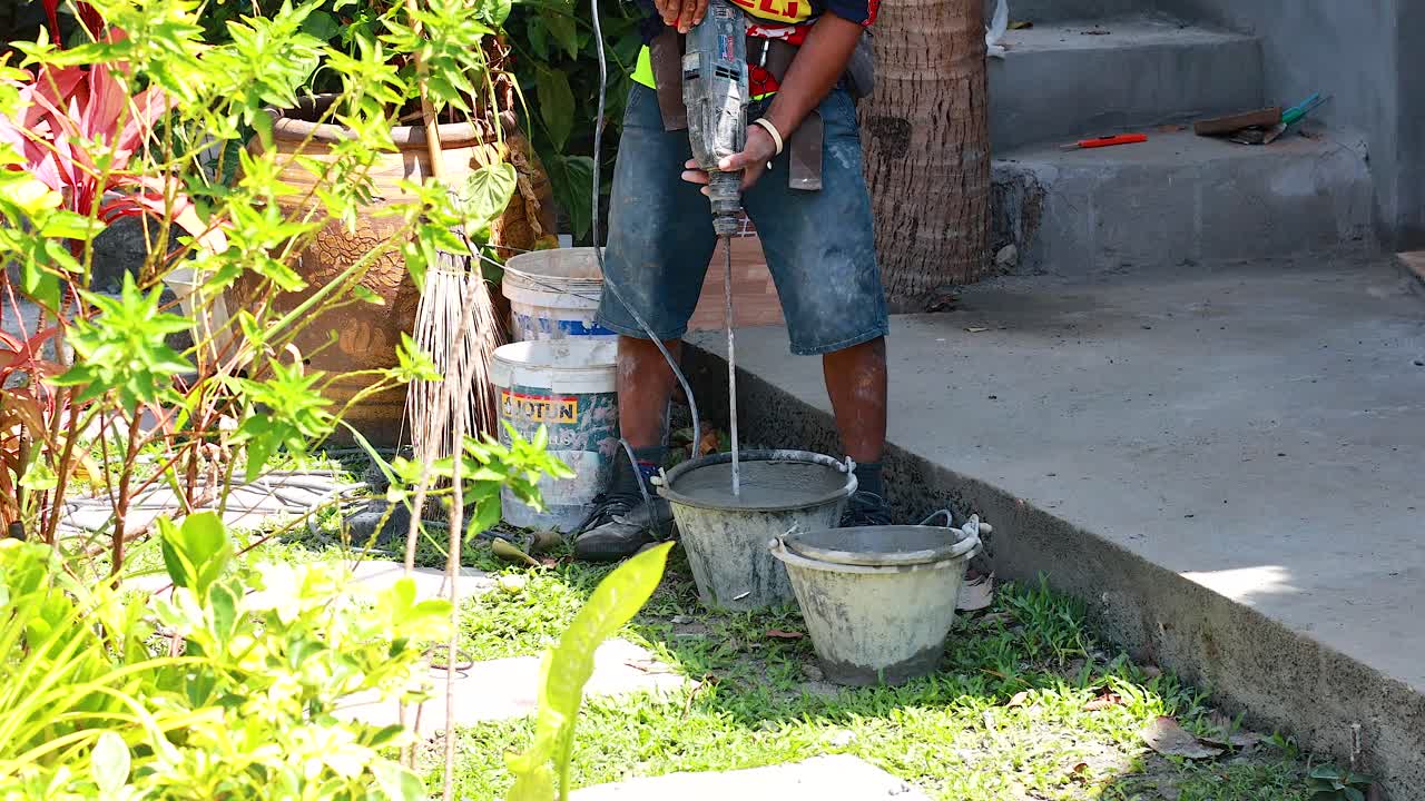A worker mixes cement in a garden setting under bright sunlight, surrounded by lush greenery and construction materials