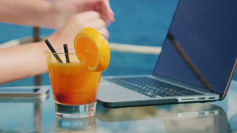 manos de mujer escribiendo en un teclado portátil portátil se encuentra en una mesa con vistas al mar cerca de un cockt