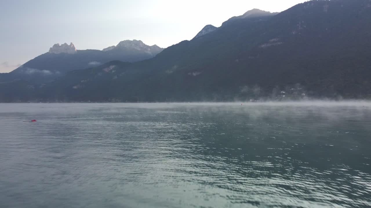 Low flying drone footage over the water of Lake Annecy on a misty morning. Some boats moored at a marina can be seen initially. Mist steams off the water as the sun rises over the mountains. France