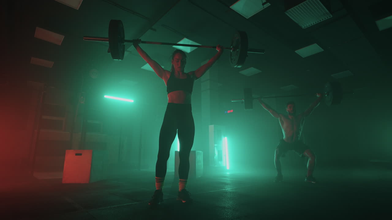 색 ⁇ 의 네온 빨간색과 파란색 빛을 입은 남자와 여자가 느린 움직임으로 머리 위에 무거운 바벨을 들어 올립니다. 무거운 무게로 힘과 힘 훈련. 운동과 강한 커플의 개념