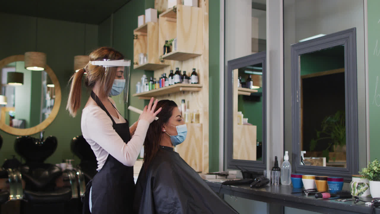 Female hairdresser wearing face cover combing hair of female customer at hair salon