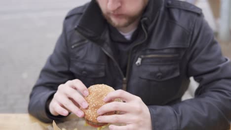 Young-man-with-beard-in-the-street-cafe-biting-tasty-big-burger-with-cheese.-French-fries-on-the-plate.-Shot-in-4k