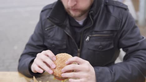 young man with beard in the street cafe biting tasty big burger with cheese. french fries on the plate. shot in 4k