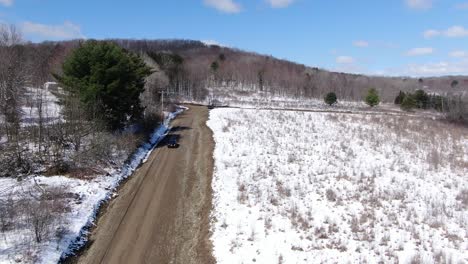 Drohnenaufnahmen-Aus-Der-Luft-Hinter-Einem-Auto,-Das-Eine-Unbefestigte-Straße-In-Der-Von-Schnee-Umgebenen-Wildnis-Hinunterfährt