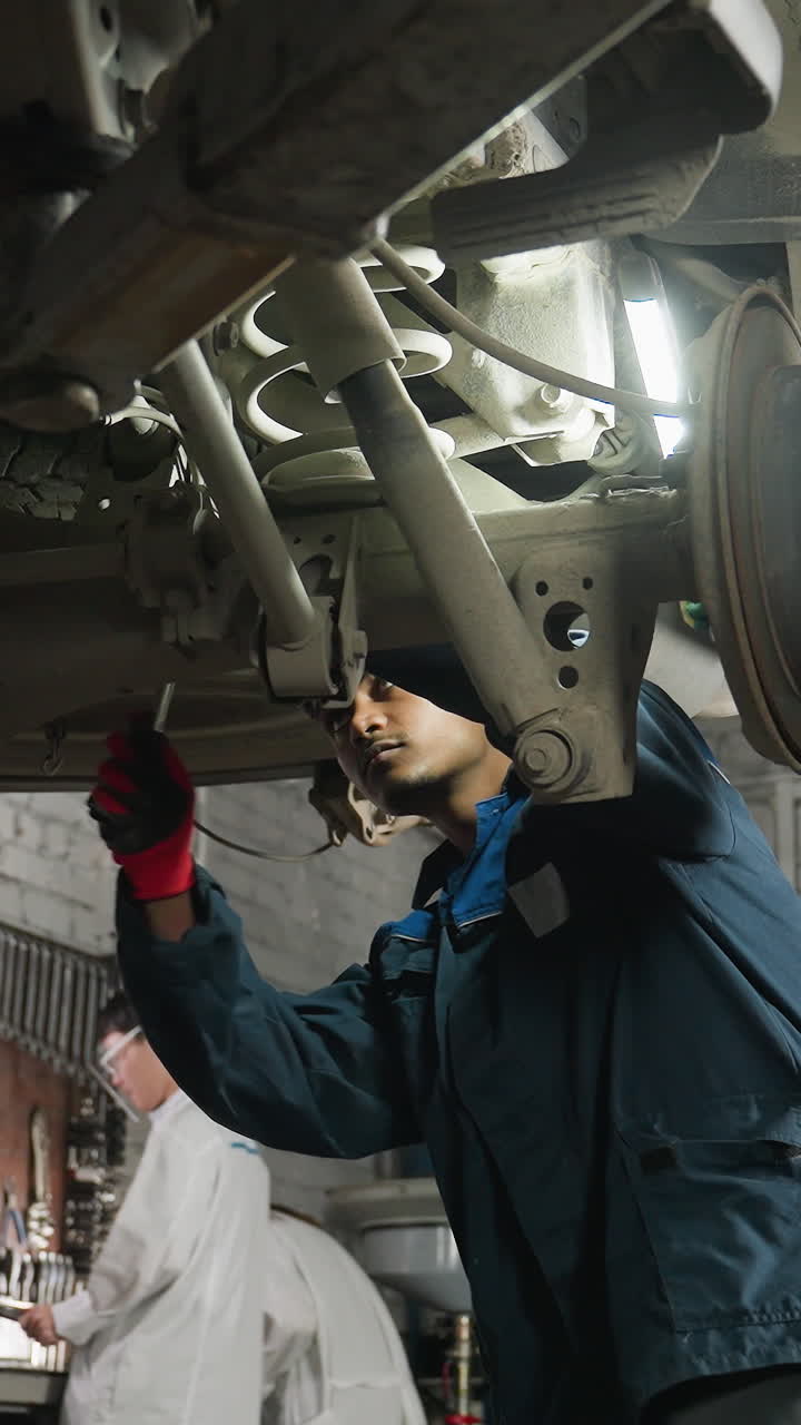 mecánico trabaja bajo el coche usando una llave mientras un colega sostiene la luz para una mejor visibilidad, dos técnicos de laboratorio se ven trabajando en el fondo en un taller mecánico