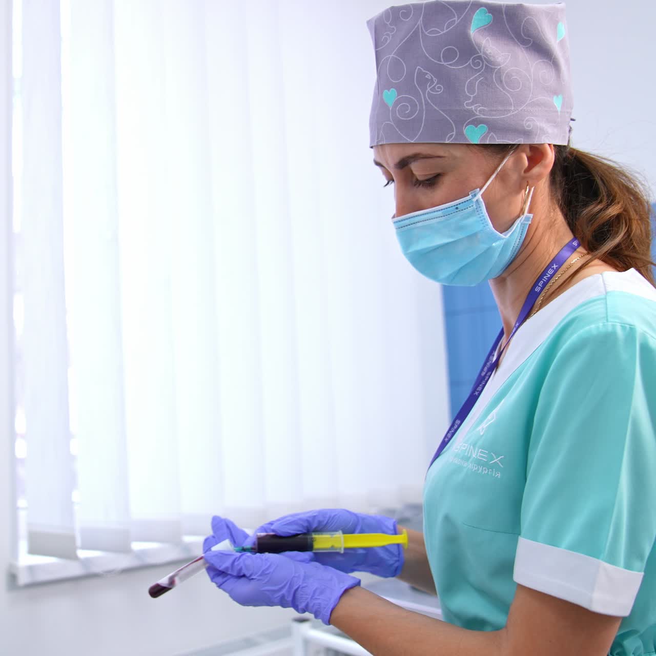 Medic in clinics wearing protective mask, gloves and cap puts blood from syringe to a test tube. Laboratory room backdrop