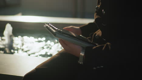 man in black coat operating tablet with one hand while seated near spring water under sunlight, reflections shimmering on water surface