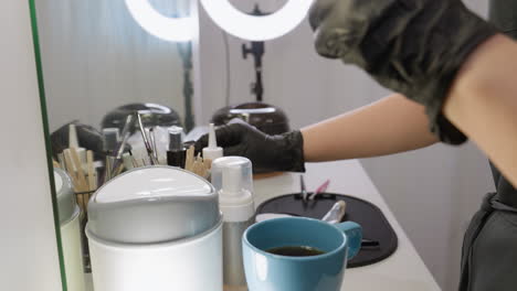 closeup view of beautician wearing gloves reaching for cotton pad and eyebrow brush on white salon table with beauty tools, cosmetic bottles, and blue mug under bright ring lights